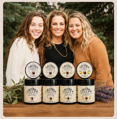 Three women standing behind six jars of YALLOW hair product on a wooden table, with greenery and lavender sprigs around the products, set against a blurred outdoor background of evergreen trees.